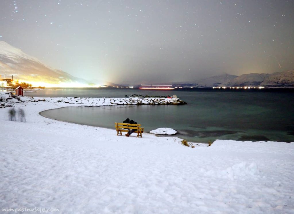 Una pareja sentada en un banco en la playa de Telegrafbukta con el cielo nocturno de fondo